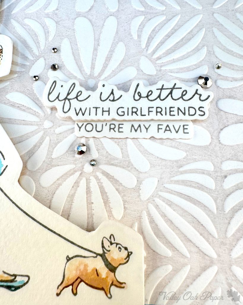 Closeup of a handmade greeting card featuring an illustration of a small fat dog.
The background is embossed with an organic, floral pattern. It's been highlighted by a light gray tint in the debossed areas.
The sentiment reads'Life is better with girlfriends. You're my fave' and is surrounded by many small silver rhinestone embellishments.