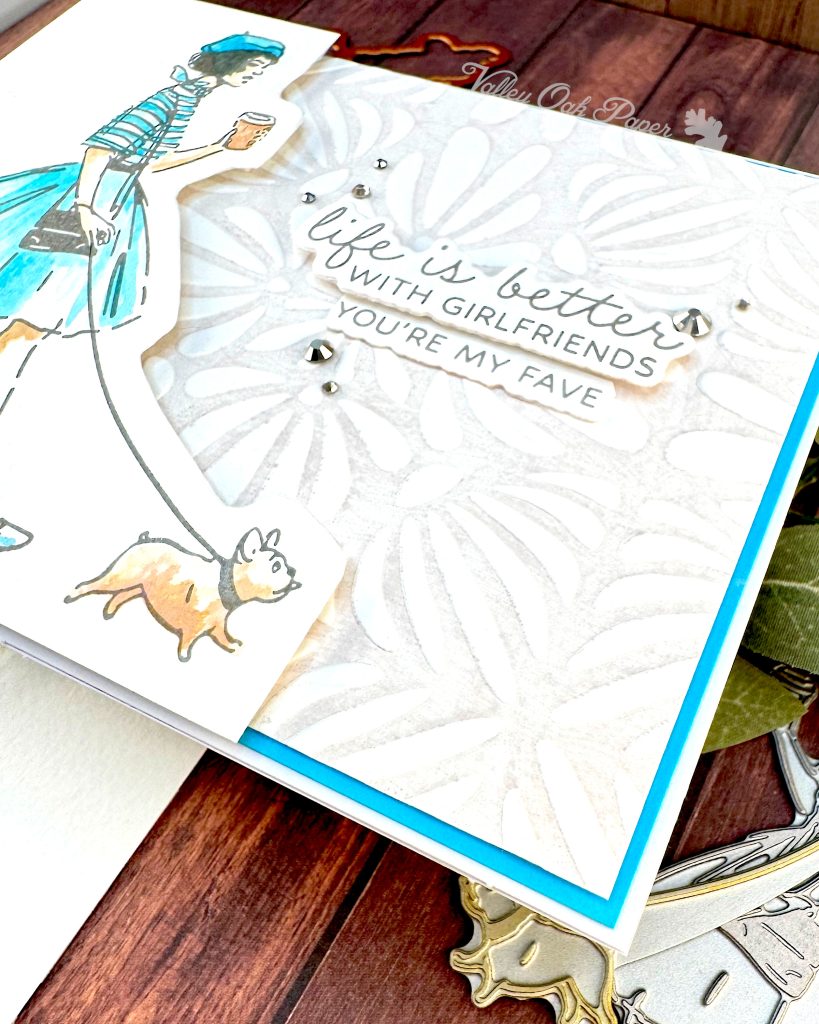 A closeup of a handmade greeting card featuring an illustration of a fashionable woman in a turquoise striped shirt, fluffy turquoise skirt, and matching beret walking a small fat dog and holding a coffee cup.
The background is embossed with an organic, floral pattern.
The sentiment reads 'Life is better with girlfriends. You're my fave' and is surrounded by small silver rhinestone embellishments.
The card is matted with a bright turquoise border. It sits on a rustic wooden background alongside crafting supplies such as BetterPress plates and dies.