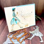 A handmade greeting card featuring an illustration of a fashionable woman in a turquoise striped shirt, fluffy turquoise skirt, and matching beret walking a small fat dog while holding a coffee cup. The background is embossed with an organic, floral pattern. The sentiment reads'Life is better with girlfriends. You're my fave' and is surrounded by small silver rhinestone embellishments. The card is matted with a bright turquoise border. It stands facing right on a rustic wooden background alongside crafting supplies such as BetterPress plates and dies.