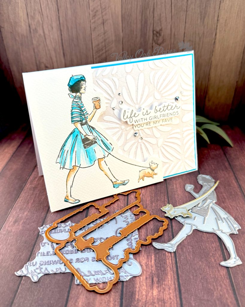 A handmade greeting card featuring an illustration of a fashionable woman in a turquoise striped shirt, fluffy turquoise skirt, and matching beret walking a small fat dog while holding a coffee cup.
The background is embossed with an organic, floral pattern.
The sentiment reads'Life is better with girlfriends. You're my fave' and is surrounded by small silver rhinestone embellishments.
The card is matted with a bright turquoise border. It stands facing right on a rustic wooden background alongside crafting supplies such as BetterPress plates and dies.
