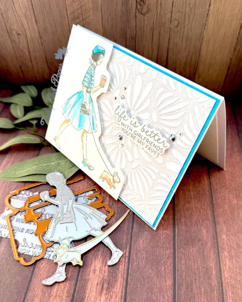 A handmade greeting card featuring an illustration of a fashionable woman in a turquoise striped shirt, fluffy turquoise skirt, and matching beret walking a small fat dog while holding a coffee cup.
The background is embossed with an organic, floral pattern.
The sentiment reads'Life is better with girlfriends. You're my fave' and is surrounded by small silver rhinestone embellishments.
The card is matted with a bright turquoise border. It stands facing left on a rustic wooden background alongside crafting supplies such as BetterPress plates and dies.