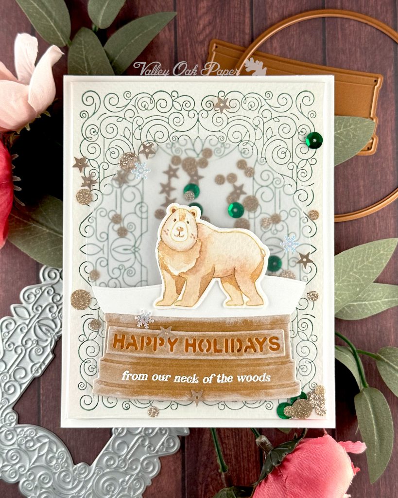 A handmade holiday card with a shaker element taking up most of the front. The shaker is filled with green sequins and champagne glitter discs. The background is an intricate pattern of symmetrical green swirls. 

On top of that sits a snow globe featuring a watercolor bear looking at the observer. The base of the snow globe reads "Happy Holidays, from our neck of the woods."

The card is photographed on a warm wooden surface decorated with roses, a letterpress plate and snow globe dies.
