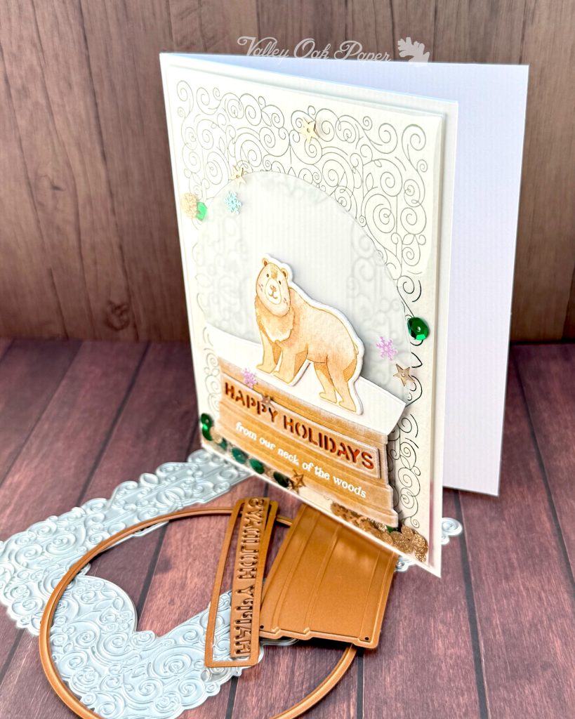 A handmade holiday card with a shaker element taking up most of the front. The shaker is filled with green sequins and champagne glitter discs. The background is an intricate pattern of symmetrical green swirls. 

On top of that sits a snow globe featuring a watercolor bear looking at the observer. The base of the snow globe reads "Happy Holidays, from our neck of the woods."

The card is photographed standing on a warm wooden surface with a letterpress plate and snow globe dies.