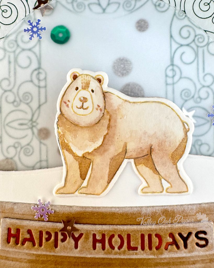 Closeup of a handmade holiday card with a shaker element taking up most of the front. The shaker is filled with green sequins and champagne glitter discs. The background is an intricate pattern of symmetrical green swirls. 

The closeup focuses on a watercolored bear. Under the bear the sentiment "Happy Holidays" is visible. 