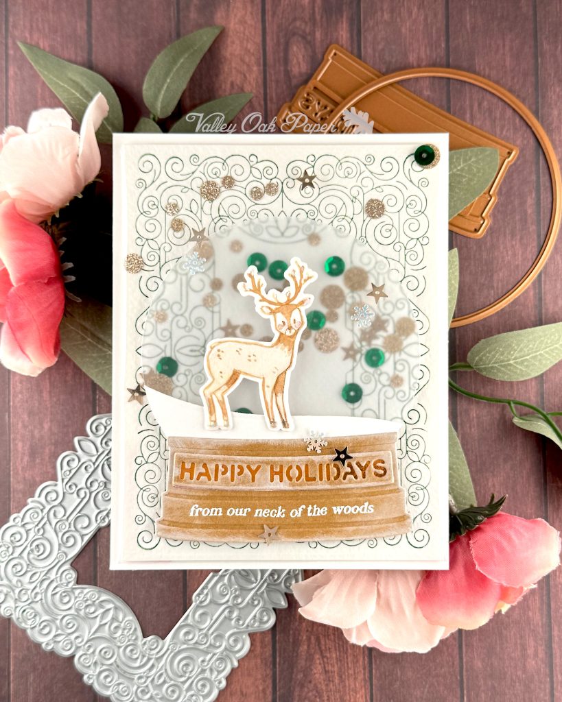 A handmade holiday card with a shaker element taking up most of the front. The shaker is filled with green sequins and champagne glitter discs. The background is an intricate pattern of symmetrical green swirls. 

On top of that sits a snow globe featuring a watercolor deer looking at the observer. The base of the snow globe reads "Happy Holidays, from our neck of the woods."

The card is photographed on a warm wooden surface decorated with roses, a letterpress plate and snow globe dies.
