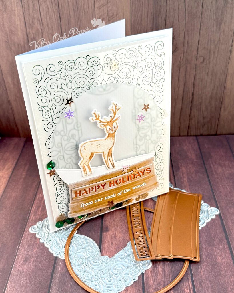 A handmade holiday card with a shaker element taking up most of the front. The shaker is filled with green sequins and champagne glitter discs. The background is an intricate pattern of symmetrical green swirls. 

On top of that sits a snow globe featuring a watercolor deer looking at the observer. The base of the snow globe reads "Happy Holidays, from our neck of the woods."

The card is photographed standing on a warm wooden surface with a letterpress plate and snow globe dies.