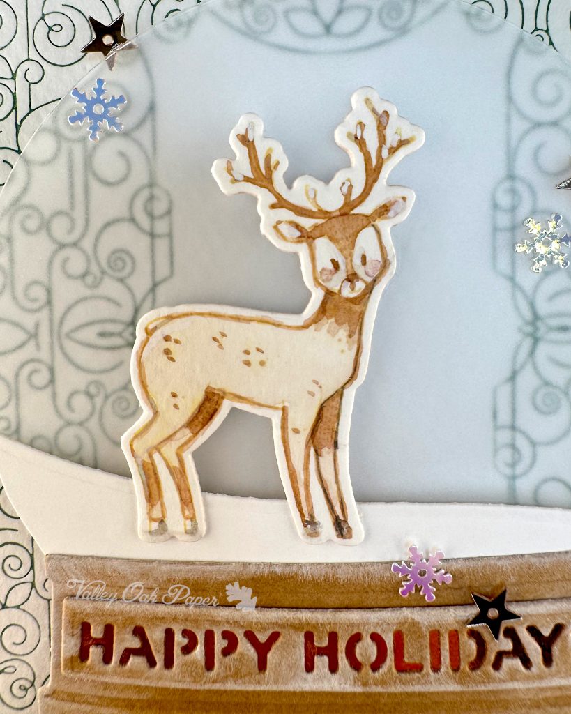 Closeup of a handmade holiday card with a shaker element taking up most of the front. The shaker is filled with green sequins and champagne glitter discs. The background is an intricate pattern of symmetrical green swirls. 

The closeup focuses on a watercolored deer with antlers. Under the bear the sentiment "Happy Holidays" is visible. 