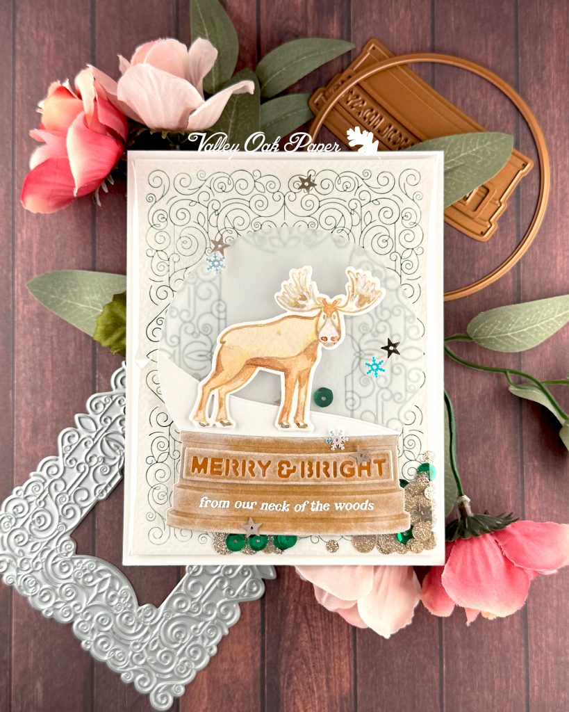 A handmade holiday card with a shaker element taking up most of the front. The shaker is filled with green sequins and champagne glitter discs. The background is an intricate pattern of symmetrical green swirls. 

On top of that sits a snow globe featuring a watercolor moose looking at the observer. The base of the snow globe reads "Merry & Bright from our neck of the woods."

The card is photographed on a warm wooden surface decorated with roses, a letterpress plate and snow globe dies.