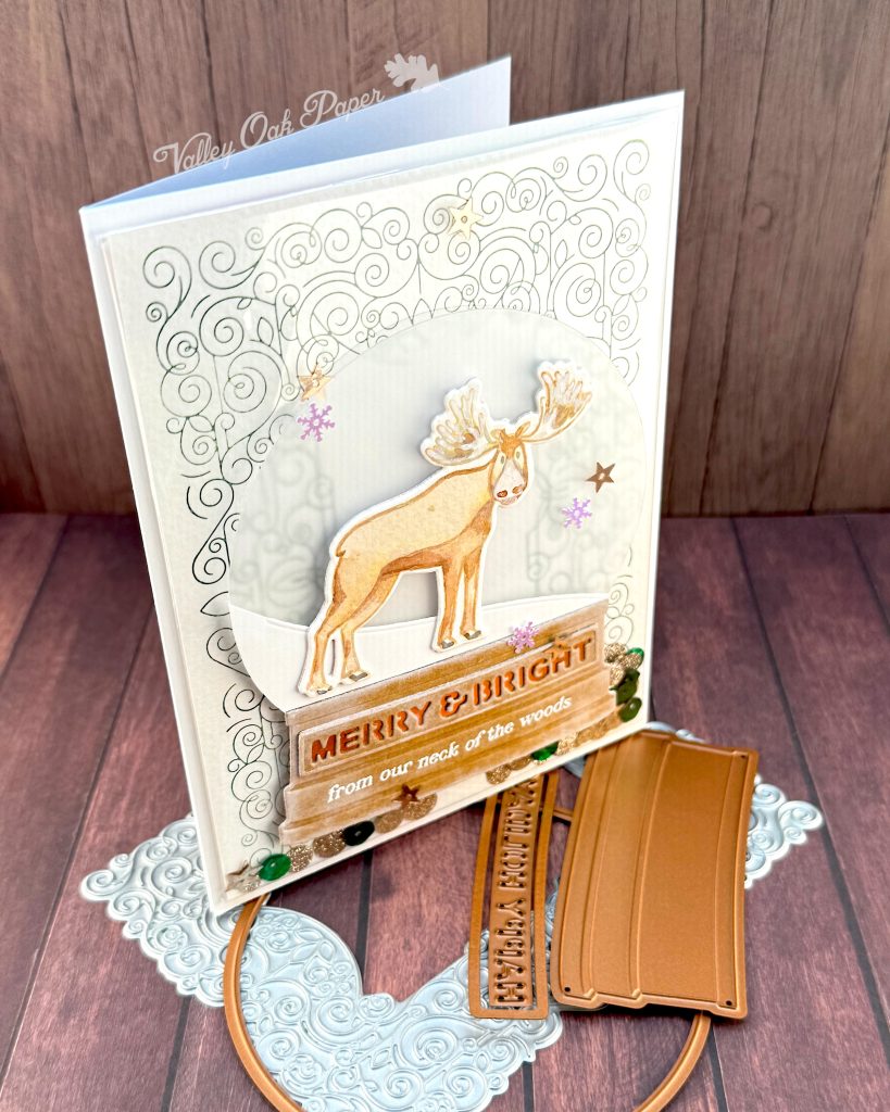 A handmade holiday card with a shaker element taking up most of the front. The shaker is filled with green sequins and champagne glitter discs. The background is an intricate pattern of symmetrical green swirls. 

On top of that sits a snow globe featuring a watercolor moose looking at the observer. The base of the snow globe reads "Merry & Bright from our neck of the woods."

The card is photographed standing on a warm wooden surface with a letterpress plate and snow globe dies.
