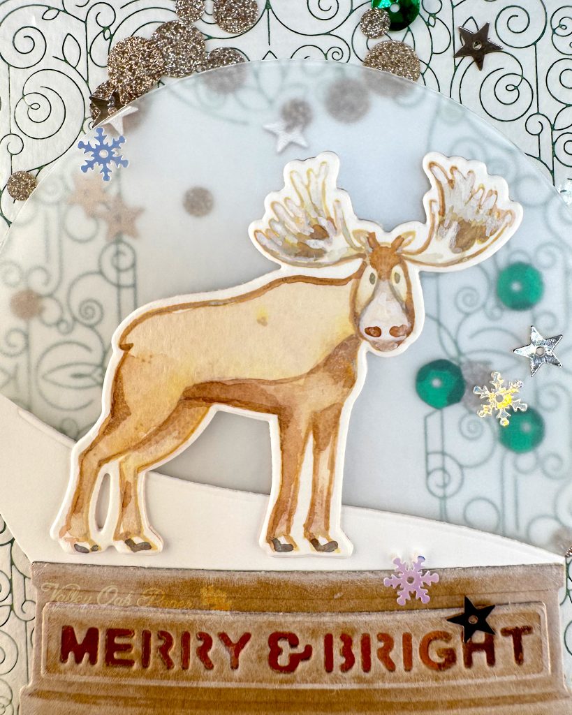 Closeup of a handmade holiday card with a shaker element taking up most of the front. The shaker is filled with green sequins and champagne glitter discs. The background is an intricate pattern of symmetrical green swirls. 

The closeup focuses on a watercolored moose with giant antlers. Under the moose the sentiment "Merry & Bright" is visible. 