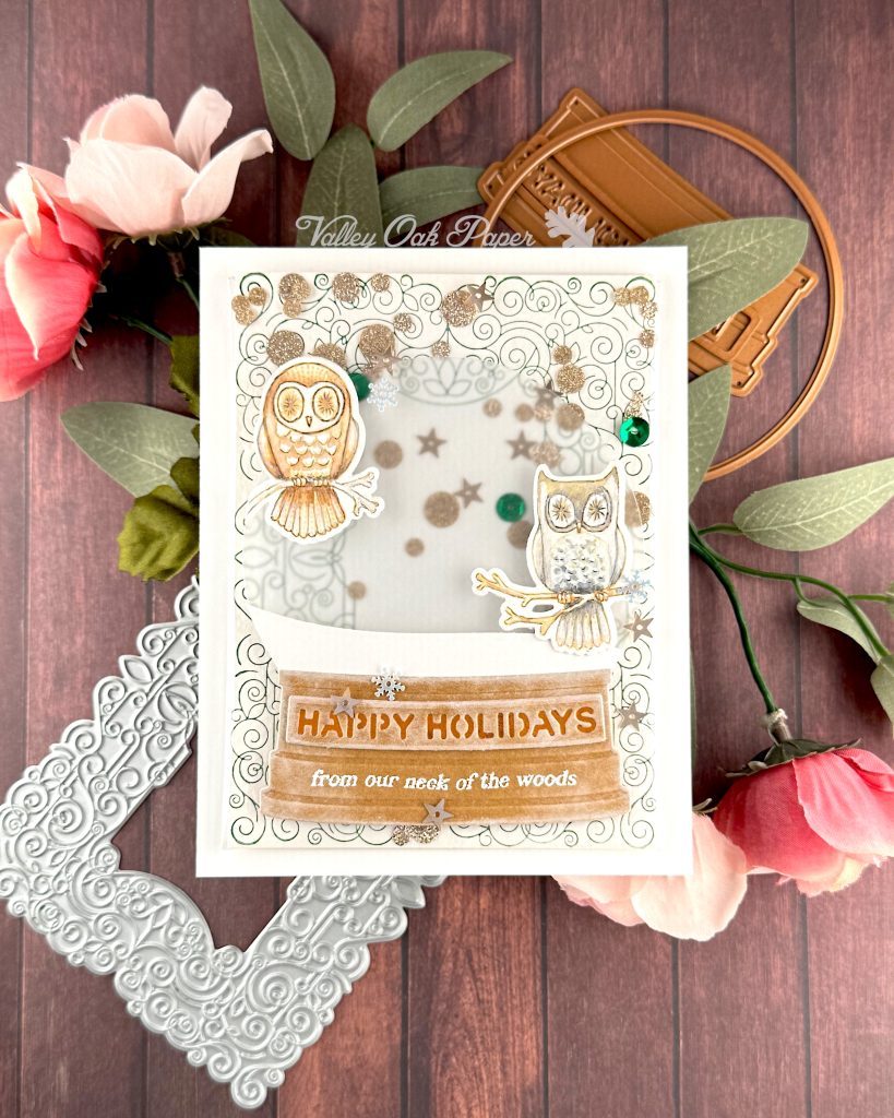 A handmade holiday card with a shaker element taking up most of the front. The shaker is filled with green sequins and champagne glitter discs. The background is an intricate pattern of symmetrical green swirls. 

On top of that sits a snow globe featuring two watercolor owls looking at the observer. The base of the snow globe reads "Happy Holidays, from our neck of the woods."

The card is photographed on a warm wooden surface decorated with roses, a letterpress plate and snow globe dies.
