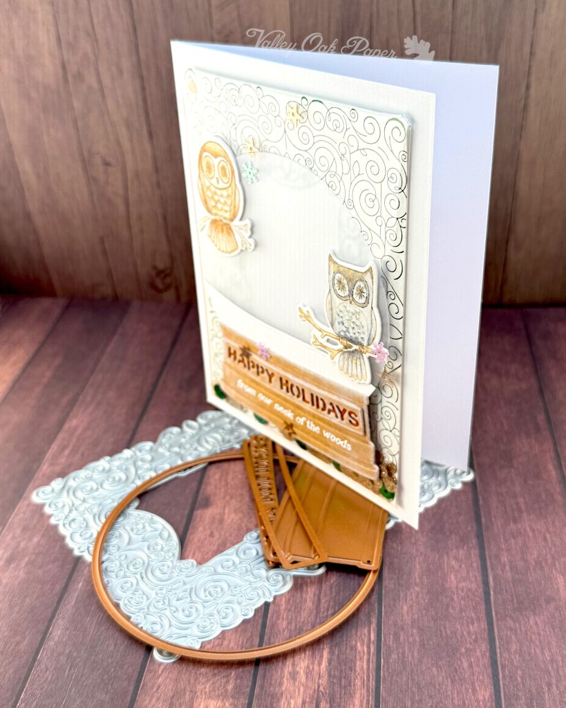 A handmade holiday card with a shaker element taking up most of the front. The shaker is filled with green sequins and champagne glitter discs. The background is an intricate pattern of symmetrical green swirls. 

On top of that sits a snow globe featuring two watercolor owls looking at the observer. The base of the snow globe reads "Happy Holidays, from our neck of the woods."

The card is photographed standing on a warm wooden surface with a letterpress plate and snow globe dies.