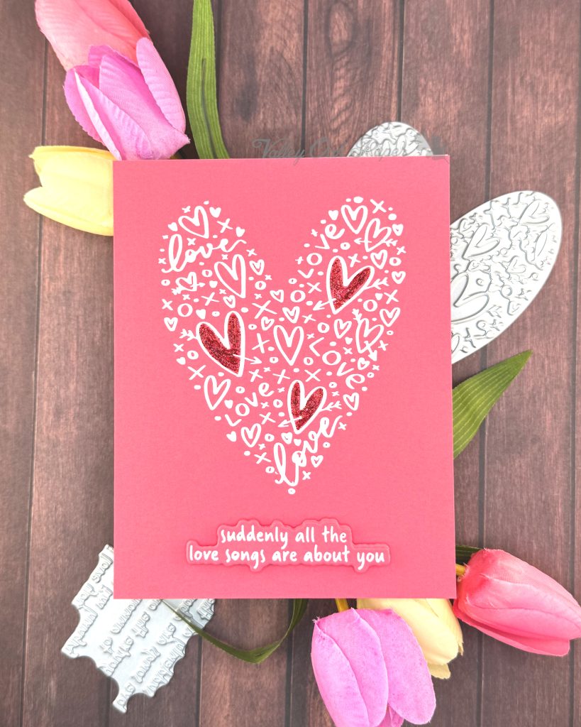 A clean and simple Valentines card in pink and white. The background is a lovely coral color. It features a heart shape made up of lots of hearts and other marks. Three of the hearts in the shape have been filled in with red glitter glue.
The sentiment sits below the heart shape. It reads "Suddenly all the love songs are about you."
The heart shape and sentiment betterpress plates peek out from under the card. So do some tulips in spring colors.
The background is a rustic wood floor.