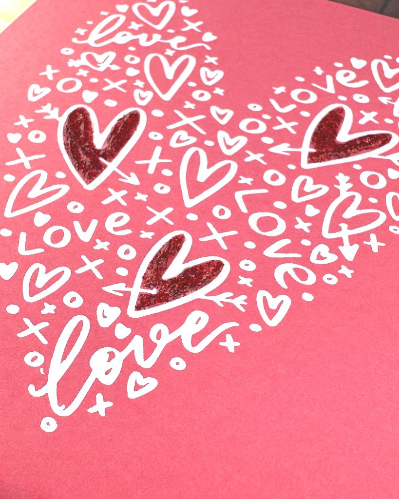 A closeup of a heart shape that consists of white hearts, x's and o's as well as the word love. Three of the largest hearts have been filled in with red glitter glue. The background is a lovely coral color.