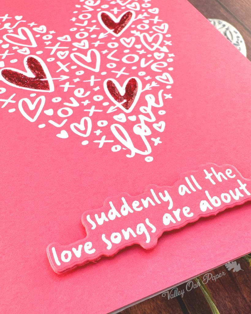A clean and simple Valentines card in pink and white. The background is a lovely coral color. It features a heart shape made up of lots of hearts and other marks. Three of the hearts in the shape have been filled in with red glitter glue.
The sentiment sits below the heart shape. It reads "Suddenly all the love songs are about you."
The photo has been taken from the bottom left corner of the card.