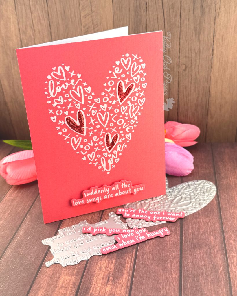 A clean and simple Valentines card in pink and white. The background is a lovely coral color. It features a heart shape made up of lots of hearts and other marks. Three of the hearts in the shape have been filled in with red glitter glue.
The sentiment sits below the heart shape. It reads "Suddenly all the love songs are about you."
The heart shape and sentiment betterpress plates peek out from under the card. So do some tulips in spring colors.
The background is a rustic wood floor. The card is standing on it, facing to the right.