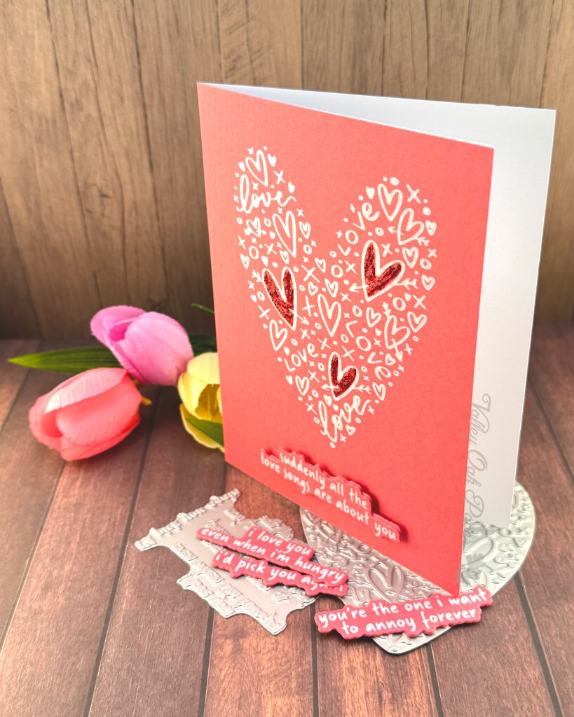 A clean and simple Valentines card in pink and white. The background is a lovely coral color. It features a heart shape made up of lots of hearts and other marks. Three of the hearts in the shape have been filled in with red glitter glue.
The sentiment sits below the heart shape. It reads "Suddenly all the love songs are about you."
The heart shape and sentiment betterpress plates peek out from under the card. So do some tulips in spring colors.
The background is a rustic wood floor. The card is standing on it, facing to the left.