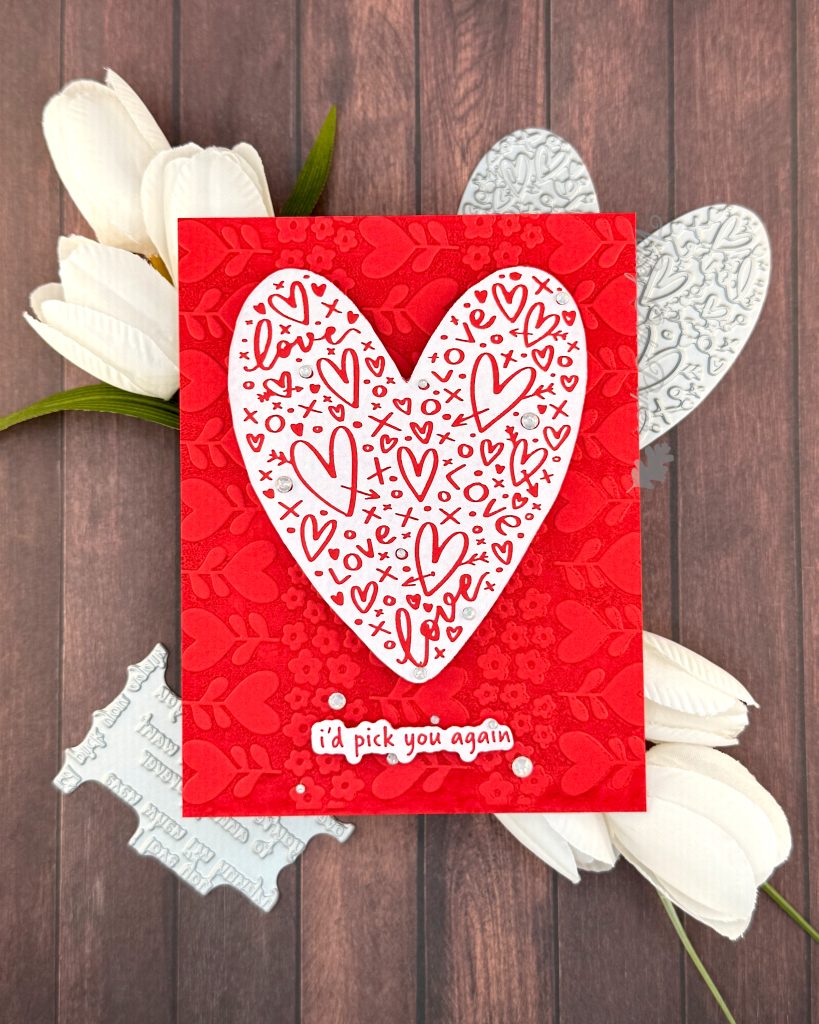 A handmade greeting card on a warm, wooden surface. The card features a white heart covered with red hearts and other markings. Some milky, white gems are scattered across its surface.
The sentiment is also white with red letters. It reads "I'd pick you again." Some white gems of various sizes are scattered around it.
The background of the card displays a red tone-on-tone texture of hearts and flowers.
The press plates used to create the heart and sentiment lie under the card. Some white tulips also peek out from under it.