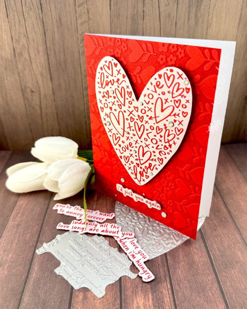 A handmade greeting card stands on a warm, wooden surface facing left. The card features a white heart covered with red hearts and other markings. Some milky, white gems are scattered across its surface.
The sentiment is also white with red letters. It reads "I'd pick you again." Some white gems of various sizes are scattered around it.
The background of the card displays a red tone-on-tone texture of hearts and flowers.
The press plates used to create the heart and sentiment lie under the card. Some white tulips and more red and white sentiments also peek out from under it.