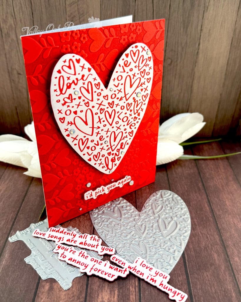 A handmade greeting card stands on a warm, wooden surface facing right. The card features a white heart covered with red hearts and other markings. Some milky, white gems are scattered across its surface.
The sentiment is also white with red letters. It reads "I'd pick you again." Some white gems of various sizes are scattered around it.
The background of the card displays a red tone-on-tone texture of hearts and flowers.
The press plates used to create the heart and sentiment lie under the card. Some white tulips and more red and white sentiments also peek out from under it.