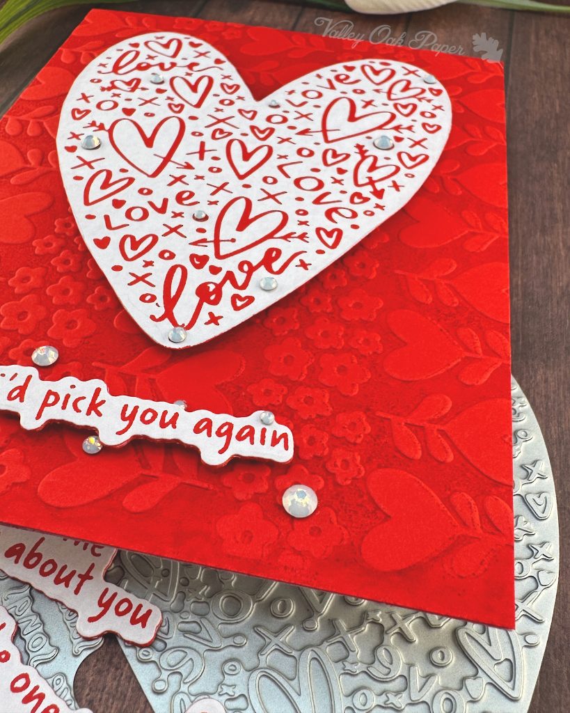 A handmade greeting card is raised off a warm, wooden surface. The card features a white heart covered with red hearts and other markings. Some milky, white gems are scattered across its surface.
The sentiment is also white with red letters. It reads "I'd pick you again." Some white gems of various sizes are scattered around it.
The background of the card displays a red tone-on-tone texture of hearts and flowers. This photo is a closeup of the bottom right corner, highlighting the texture.
The press plates used to create the heart and sentiment lie under the card. Some white tulips and more red and white sentiments also peek out from under it.
