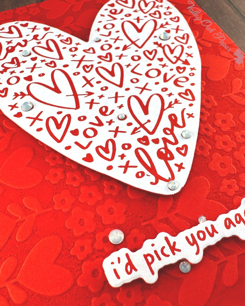 A handmade greeting card. The card features a white heart covered with red hearts and other markings. Some milky, white gems are scattered across its surface.
The sentiment is also white with red letters. It reads "I'd pick you again." Some white gems of various sizes are scattered around it.
The background of the card displays a red tone-on-tone texture of hearts and flowers. This photo is a closeup of the bottom right corner, highlighting the texture and the crisp white sentiment.
The press plates used to create the heart and sentiment lie under the card. Some white tulips and more red and white sentiments also peek out from under it.