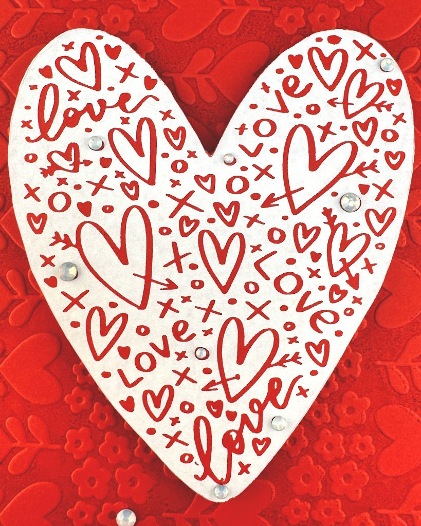 A handmade greeting card features a white heart covered with red hearts and other markings. Some milky, white gems are scattered across its surface.
We can't see the card sentiment but the background of the card displays a red tone-on-tone texture of hearts and flowers.