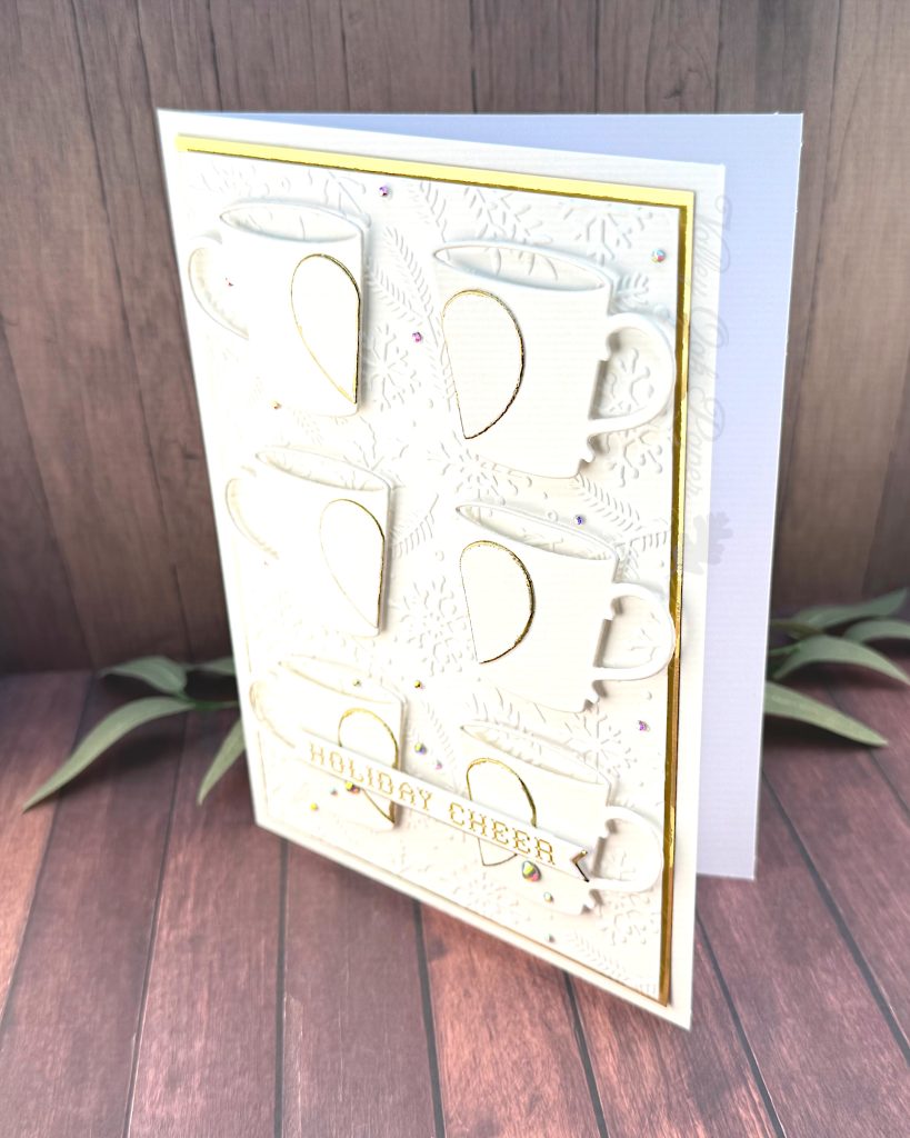 A handmade greeting card in white and gold tones stands on a warm, wooden background with some greenery, facing left.
The card is a large format 5" by 7". It has a thin gold foil matte and the warm white background is textured with snowflakes, pine branches and holly.
The card features six mugs arranged in a 2 by 3 grid. Each mug is decorated by half a heart outlined in gold.
On top of the bottom mugs, sits the sentiment on a banner. It reads "Holiday cheer" in a gold cross stitch font.