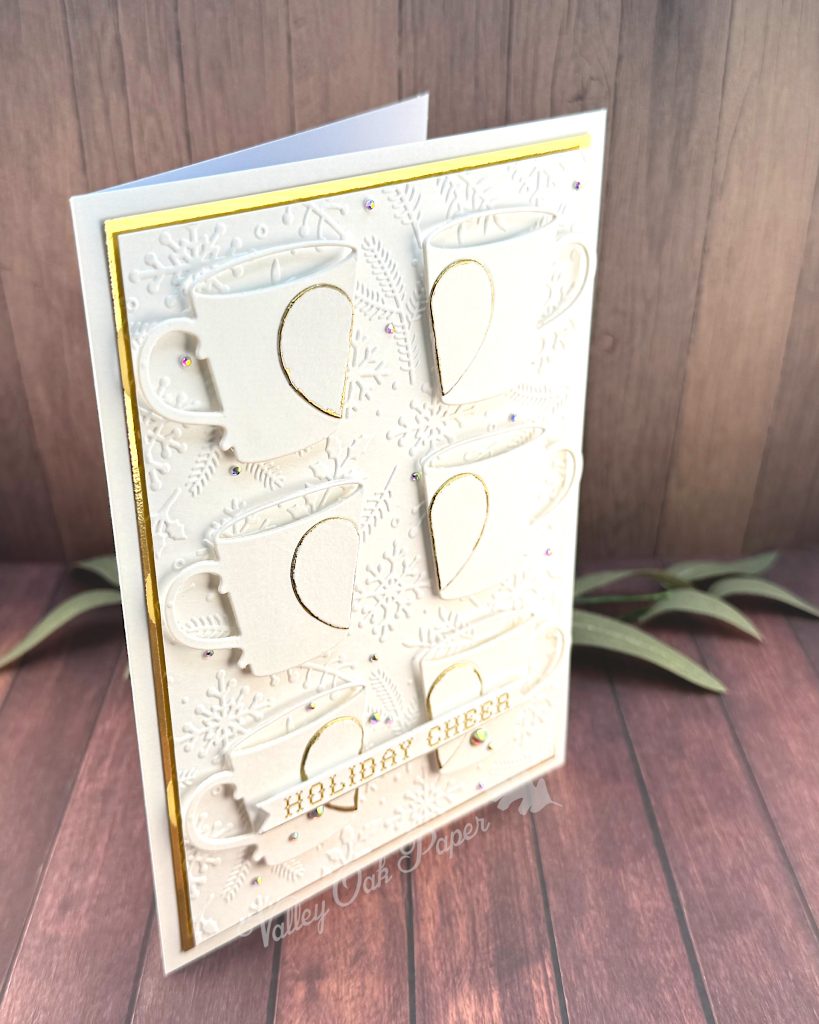 A handmade greeting card in white and gold tones stands on a warm, wooden background with some greenery, facing right.
The card is a large format 5" by 7". It has a thin gold foil matte and the warm white background is textured with snowflakes, pine branches and holly.
The card features six mugs arranged in a 2 by 3 grid. Each mug is decorated by half a heart outlined in gold.
On top of the bottom mugs, sits the sentiment on a banner. It reads "Holiday cheer" in a gold cross stitch font.