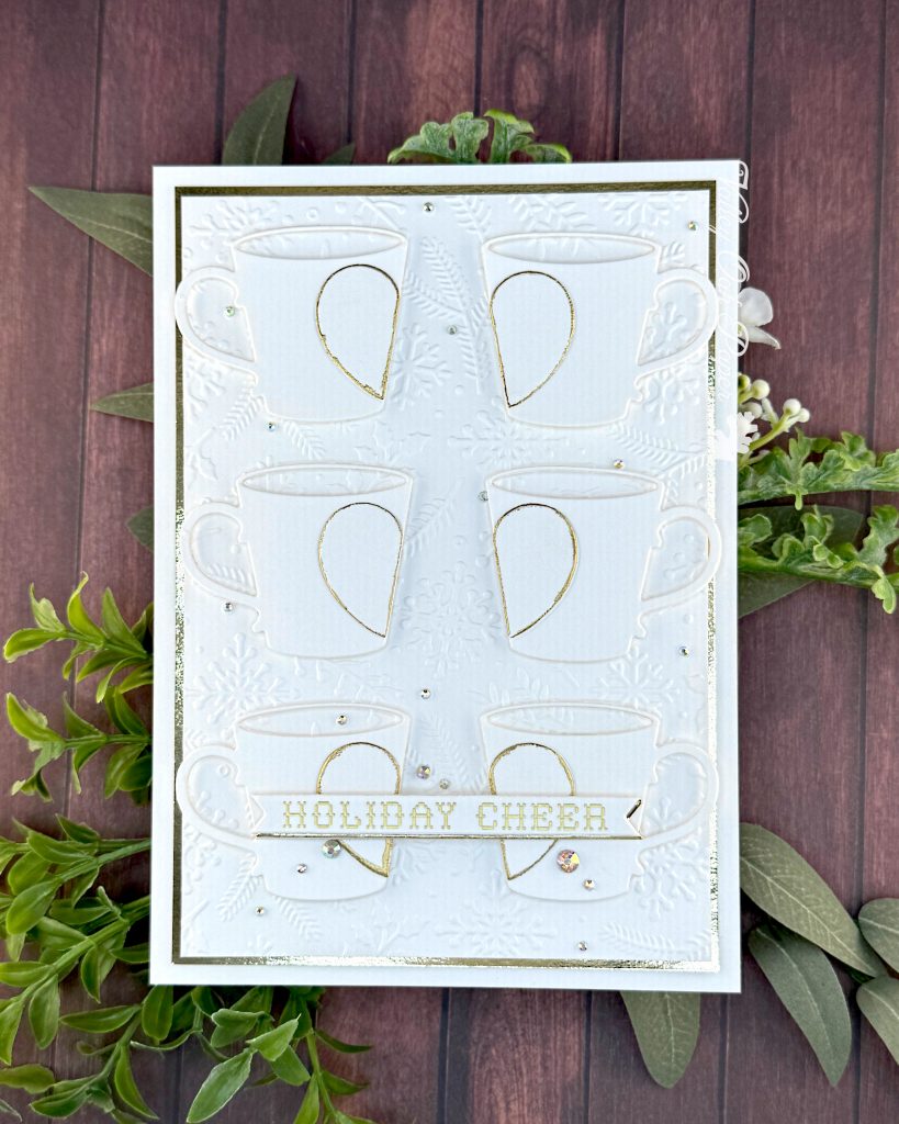A handmade greeting card in white and gold tones lies on a warm, wooden background with some greenery.
The card is a large format 5" by 7". It has a thin gold foil matte and the warm white background is textured with snowflakes, pine branches and holly.
The card features six mugs arranged in a 2 by 3 grid. Each mug is decorated by half a heart outlined in gold.
On top of the bottom mugs, sits the sentiment on a banner. It reads "Holiday cheer" in a cross stitch font.