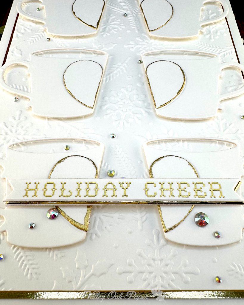 Closeup of a handmade greeting card in white and gold tones.
It has a thin gold foil matte and the warm white background is textured with snowflakes, pine branches and holly.
The card features six mugs arranged in a grid. Each mug is decorated by half a heart outlined in gold.
The closeup focuses on the bottom two mugs and the sentiment. In this shot, we see that the white sentiment banner sits on top of a slightly-offset gold banner.