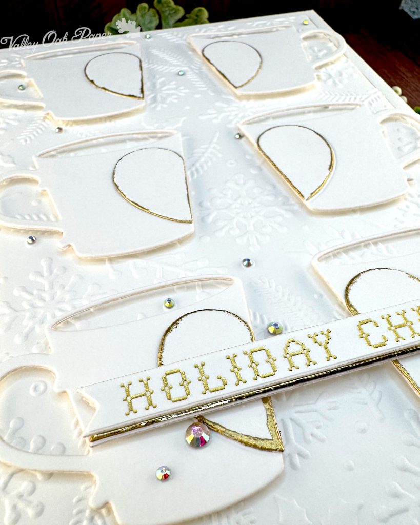 Closeup of a handmade greeting card in white and gold tones. It's lying on a warm, wooden background with some greenery.
The card is a large format 5" by 7". It has a thin gold foil matte and the warm white background is textured with snowflakes, pine branches and holly.
The card features six mugs arranged in a 2 by 3 grid. Each mug is decorated by half a heart outlined in gold.
On top of the bottom mugs, sits the sentiment on a banner. It reads "Holiday cheer" in a gold cross stitch font.
The card has been photographed from the bottom left corner. The angle emphasizes the gold foiled sentiment.