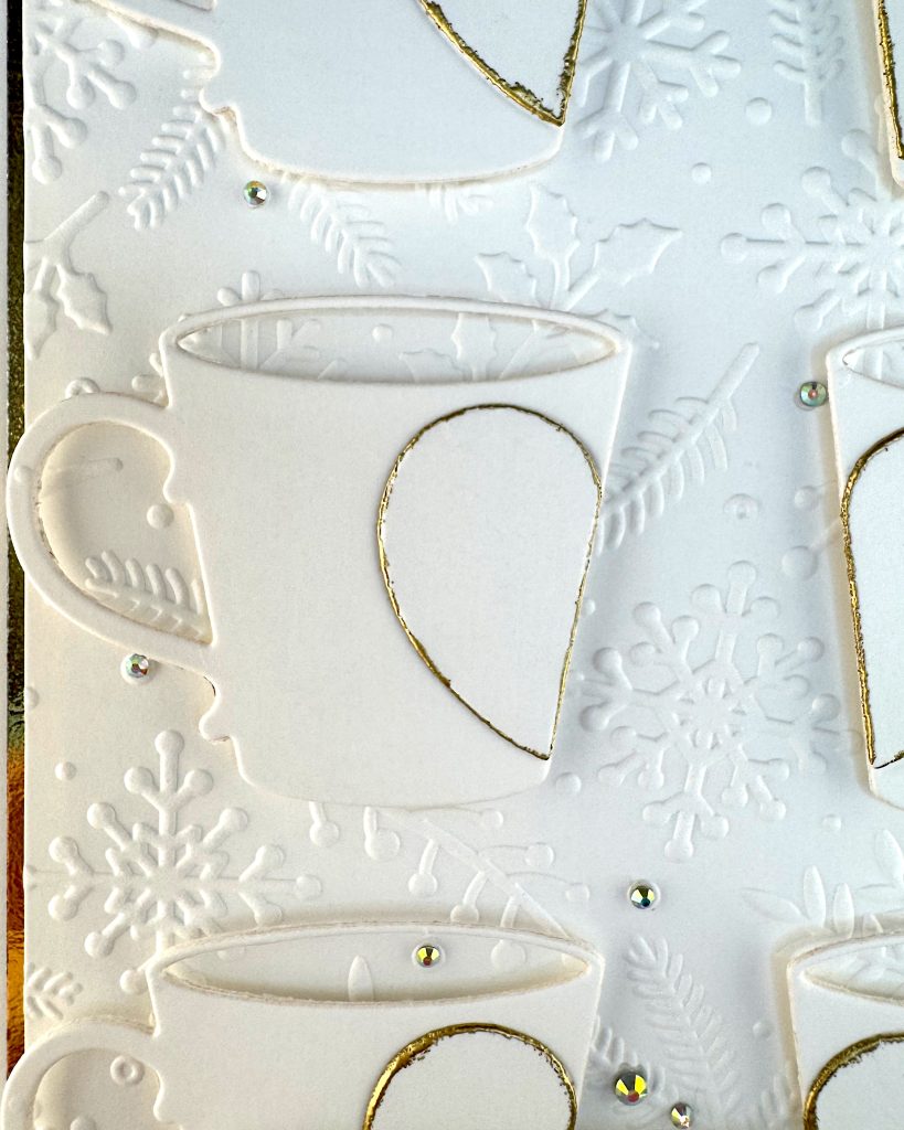 Closeup of a handmade greeting card in white and gold tones.
It has a thin gold foil matte and the warm white background is textured with snowflakes, pine branches and holly.
The card features mugs arranged in a grid. Each mug is decorated by half a heart outlined in gold.
The closeup focuses on one of the mugs. The many gold gems scattered across the surface of the card are highlighted by this crop.