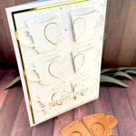 A handmade greeting card in white and gold tones stands on a warm, wooden background with some greenery, facing right. The card is a large format 5" by 7". It has a thin gold foil matte and the warm white background is textured with snowflakes, pine branches and holly. The card features six mugs arranged in a 2 by 3 grid. Each mug is decorated by half a heart outlined in gold. On top of the bottom two mugs, sits the sentiment on a banner. It reads "Holiday cheer" in a gold cross stitch font. In front of the card the dies used to create the mugs and hearts lie.